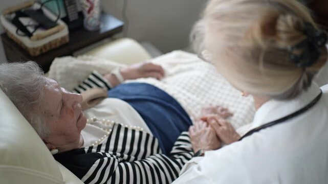A mature woman in casual attire sits comfortably at home, holding hands with her caregiver during a warm visit in the afternoon light.