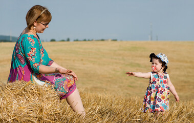 Happy mother and toddler girl playing in a summer field with haystacks. Candid outdoor moment, colorful clothing, parenting, and family bonding concept.
