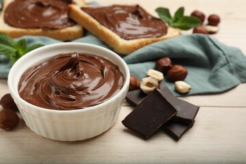 Chocolate hazelnut spread in bowl, nuts, toasts, pieces of chocolate and mint on wooden table, closeup