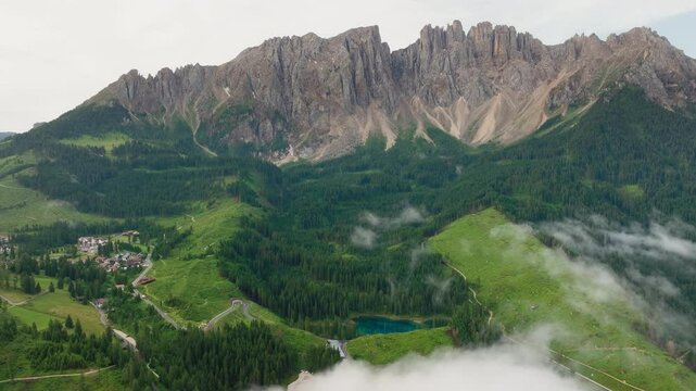 Wide aerial shot of Lago di Carezza in the Italian Alps. Lush springtime forests, tranquil lake waters and the iconic mountain in the background