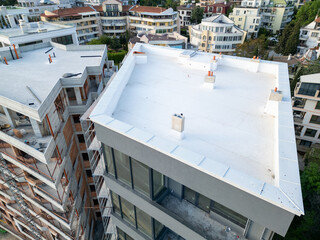 Aerial view of modern residential buildings under construction, featuring flat white rooftops, scaffolding, and unfinished facades. Surrounding neighborhood with completed homes in background