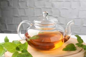 Aromatic lemon balm tea in glass teapot and fresh leaves on table against light background, closeup