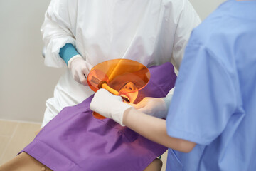 Closeup of curing light being used in dental treatment on adult asian female patient as dental assistant holds orange curing shield to protect eyes during cavity filling session in clinical room.