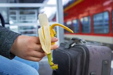 A peeled banana held by a hand of a traveller sitting on the bench by the suitcase, waiting for the...