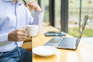 A man is sitting at a table with a laptop and a cup of coffee. He is holding the cup with his right hand and the laptop with his left. The scene suggests a relaxed and comfortable atmosphere