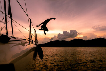 Person leaps off a sailboat at sunset over calm water with clouds and hills in the background