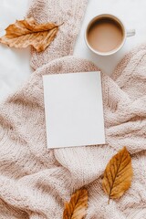 A5 blank card mockup, beige and light pink tones, cozy knitted blanket with a coffee cup nearby, dried leaves around the frame, top view, white background