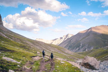 Obraz premium Photo from the back of a couple of hikers walking on green grasslands in the Tianshan Mountains in Xinjiang, China. The evening sunlight casts a soft glow on the peaks on a clear day with copy space.