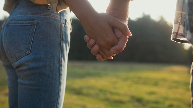 Close camera view of husband and wife holding hands in middle of green field. Sun shining bright in background. Sunset. Loving couple. Boyfriend and girlfriend. Green trees in background.
