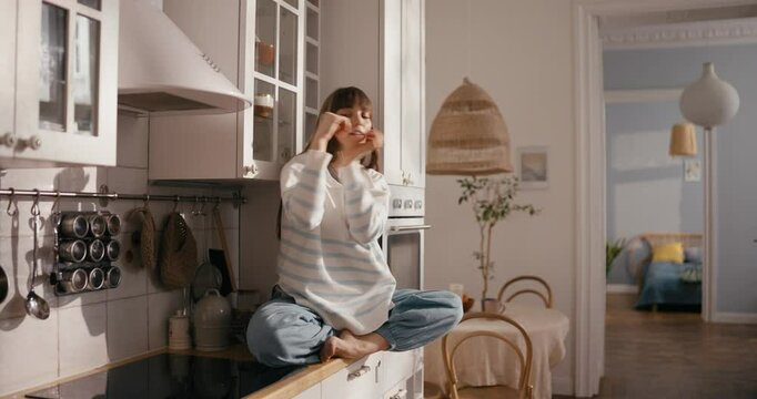 Girl in casual clothes sits on countertop and dances waving and hand dance during self-expression in kitchen of cozy apartment. Girl dancing in the kitchen