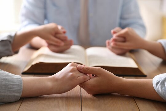 Family with Bible praying at wooden table indoors, closeup