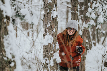 A cheerful woman wearing a hat and jacket stands among snowy trees in a lush winter forest, smiling...