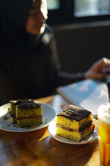 Slices of cheese chocolate cake and matcha chocolate on a plate with a blurred background of a woman in a hijab reading a book