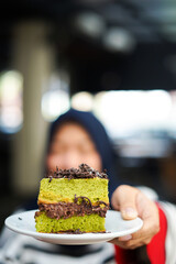 A slice of green tea cake with chocolate filling, sprinkled with grated chocolate, on a small white plate served by hand