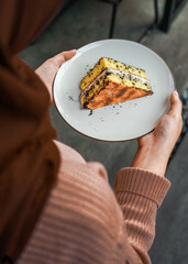 Hands holding a white plate with a tempting piece of layered cake, with chocolate crumbs and cream filling. A delicious dessert