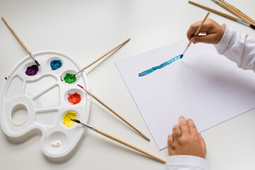 Child painting with brushes and color palette on white surface