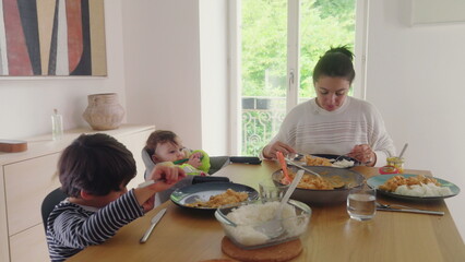 Mother feeding baby at the dining table while older child eats, family mealtime interaction, peaceful indoor environment, mother multitasking, baby in high chair, family care