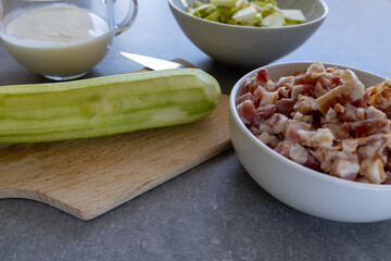 Close-up of a peeled zucchini on a wooden cutting board next to a bowl of chopped bacon, with cream and sliced zucchini in the background. 