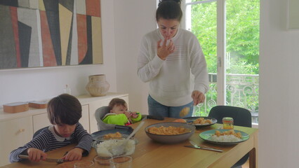 Mother serving food at the dining table while older boy and baby sit, family meal preparation and gathering in a cozy home