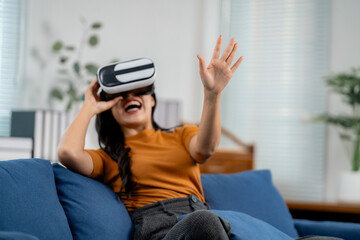 Fototapeta premium Young woman wearing virtual reality headset and interacting with virtual world while sitting on comfortable sofa at home
