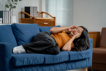 Stressed asian woman having migraine and touching her temples while lying on sofa in living room