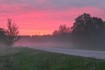 A serene rural road at sunrise with a misty foreground, silhouetted trees, and a pink-orange sky full of scattered clouds.