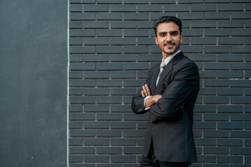 Confident young businessman standing in a formal black suit and tie against a dark brick wall, posing with a professional and self assured expression.