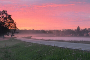 A serene rural road at sunrise with a misty foreground, silhouetted trees, and a pink-orange sky full of scattered clouds.