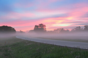 A serene rural road at sunrise with a misty foreground, silhouetted trees, and a pink-orange sky full of scattered clouds.
