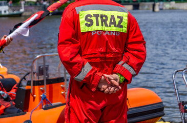 Polish water rescue firefighter in red uniform with "STRAŻ POLAND" label near an orange rescue boat by the water © Adam