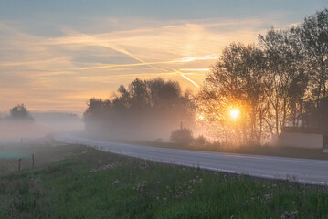 A glowing golden sunrise lights up a foggy rural road with trees silhouetted against the mist and a vivid yellow-orange sky.