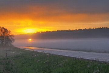 A glowing golden sunrise lights up a foggy rural road with trees silhouetted against the mist and a vivid yellow-orange sky.