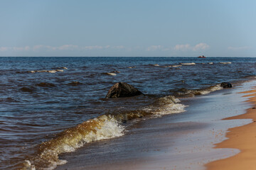 A dark rock partially submerged in sea water with small waves crashing on a sandy beach under sunlight.