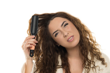 Woman with curly brown hair in white bathrobe struggling with hair brush caught in tangled hair against white background, frustrated expression, styling challenge