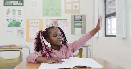 In classroom, African American girl raising hand while writing in notebook, at school - Powered by Adobe
