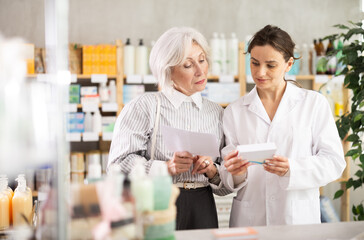 Mature female client purchases pills to treat chronic disease. Woman pharmacist in background offers product, promotes product of European brand, recommend safe and effective pills.