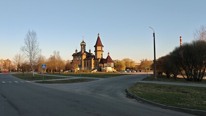 Obraz premium A wooden Orthodox church with domes and crosses stands on a stone foundation. Nearby trees, lawn, sidewalks, lampposts, houses and a factory chimney. The spring sunset sun illuminates the surroundings