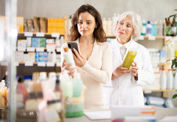 Near display case in pharmacy, woman scans barcode on shampoo packaging, reads user manual and consulting with senior woman pharmacist about using cosmetic care product
