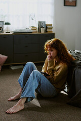 Woman sitting on floor beside bed, resting head on hand, appearing deep in thought. Room featuring...