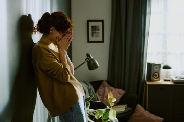 Red-haired woman standing against wall in dimly lit room covering her face, appearing distraught....