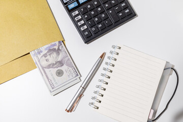 A tabletop arrangement featuring a stack of cash, a blank notepad, and a calculator—perfectly...
