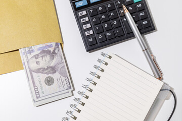 A tabletop arrangement featuring a stack of cash, a blank notepad, and a calculator—perfectly...