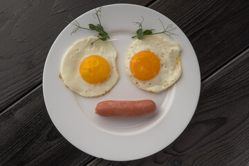Fried eggs with bright yolks and a sausage on a white plate, decorated with pea shoots, dark...