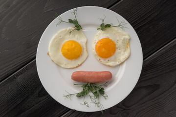 Two sunny side up eggs and a sausage on a white plate, with a dark wooden backdrop.