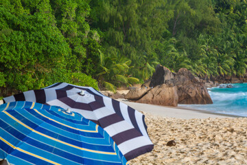 Umbrella on the sandy beach. Blue Indian Ocean, blue sky with clouds, green rocky hills and palm leaves around, ANSE INTENDANCE, MAHE ISLAND, THE SEYCHELLES