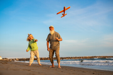 Grandfather and grandson joyfully watching toy airplane fly above beach, enjoying playful moment together near sea on sunny day.