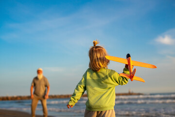 Little boy holding an orange toy airplane on the beach while looking at his grandfather in the background, enjoying playful family time by the sea during golden hour.