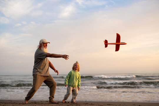 Elderly man launching a toy airplane on the beach while his cheerful blond grandson watches, both enjoying playful moments by the ocean at sunset.