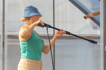 A close-up view of a woman using a pressure washer to clean the greenhouse walls. The image focuses on greenhouse maintenance and agricultural care.