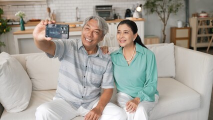 Obraz premium Happy grandfather and grandmother making video call with their son at home. Elder man holding smart phone and sitting at sofa while old woman greeting with cheerful vibe at modern house. Myrmidon.
